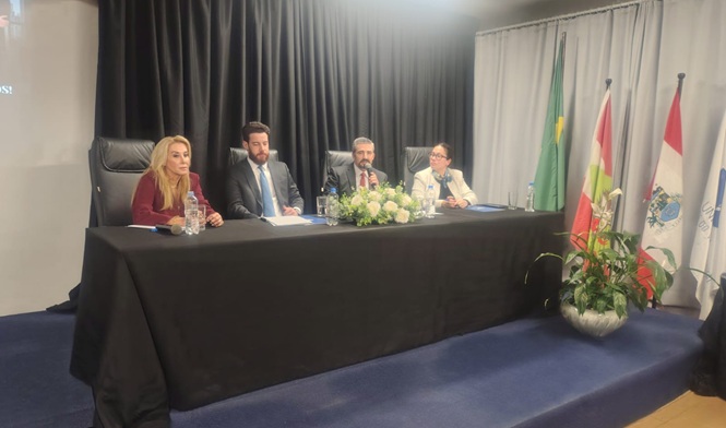 Foto horizontal: mesa de autoridades em evento institucional, participantes sentados com bandeiras ao fundo, clima de debate e cooperação.