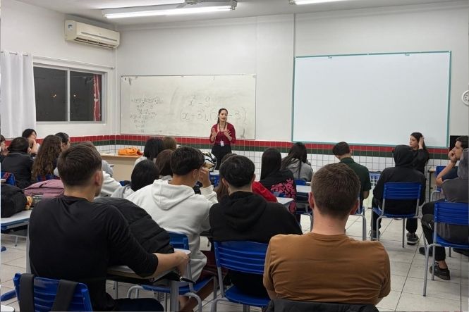 Foto horizontal: mulher conduz apresentação diante de estudantes sentados em sala de aula, com quadros brancos ao fundo.