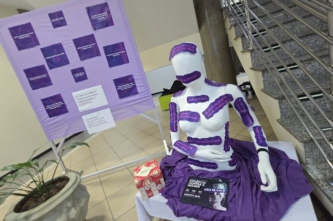 Foto horizontal: manequim com mensagens em tons de roxo integra instalação de conscientização, ao lado de painel informativo, em área interna próxima a uma escadaria.