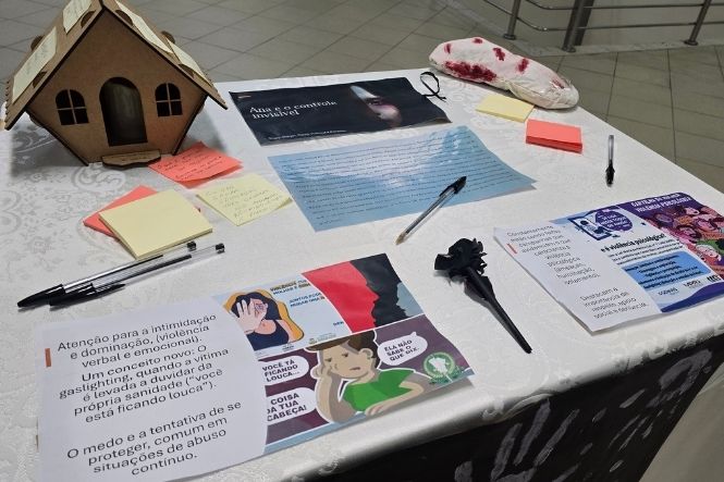 Foto horizontal: mesa reúne materiais informativos sobre violência contra a mulher, com cartilhas, mensagens, canetas e objetos que integram ação de conscientização.