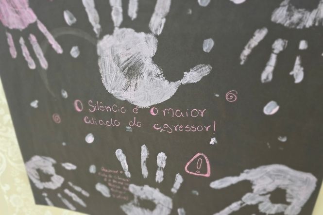 Foto horizontal: painel exibe marcas de mãos e a frase “O silêncio é o maior aliado do agressor!”, em ação de conscientização contra a violência.