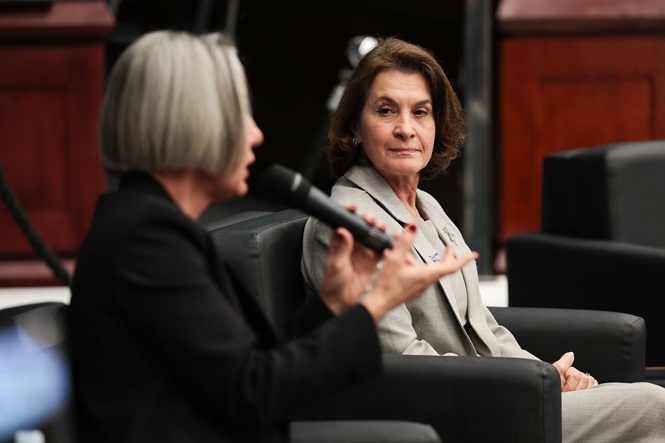 Foto horizontal: Vera Iaconelli fala ao microfone enquanto outra participante acompanha sua fala durante o evento.