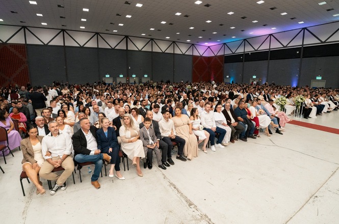 Foto horizontal: grande público reunido em cerimônia de casamento coletivo, casais sentados em fileiras, ambiente amplo e festivo, tons claros.
