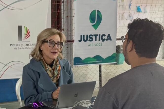 Foto horizontal: atendimento do projeto Justiça até Você, profissional utiliza notebook para orientar cidadão, banner institucional ao fundo, ambiente comunitário, clima de acolhimento.