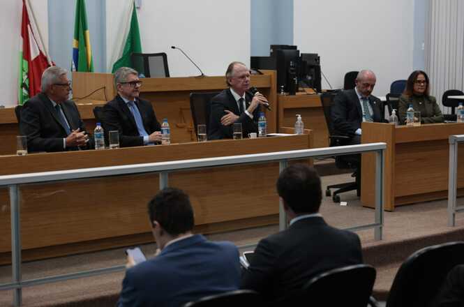 Foto horizontal: sessão institucional com autoridades à mesa, desembargador Rubens Schulz fala ao microfone, bandeiras ao fundo, ambiente formal.