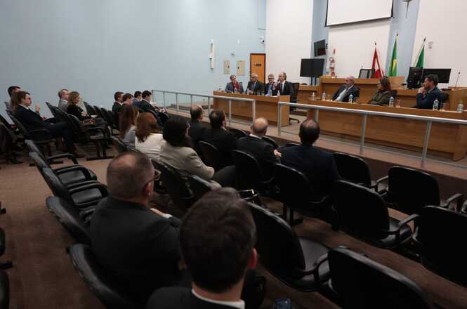 Foto horizontal: sessão institucional com autoridades à mesa e público presente, plenário parcialmente ocupado, ambiente formal, clima de escuta e participação.