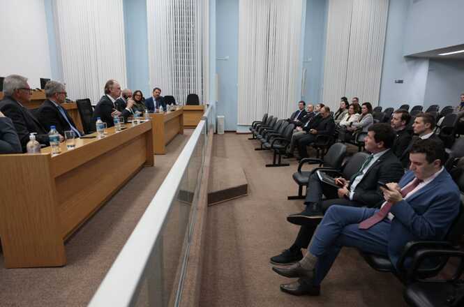 Foto horizontal: sessão institucional com autoridades à mesa e público presente, debate em andamento, ambiente formal, clima de escuta e participação.