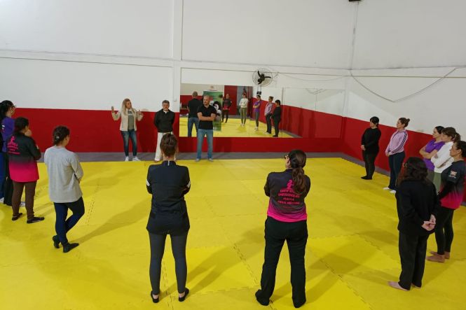 Foto horizontal: grupo participa de dinâmica em espaço coletivo, pessoas em círculo sobre tatame, clima de escuta, acolhimento e fortalecimento coletivo.