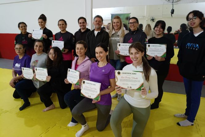 Foto horizontal: grupo de mulheres exibe certificados em atividade coletiva, ambiente de integração, clima de conquista, fortalecimento e valorização feminina.