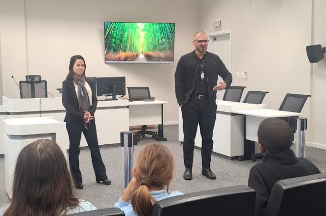 Foto horizontal: servidores recebem grupo de estudantes em sala de audiências e apresentam o funcionamento do Poder Judiciário durante visita guiada ao fórum.