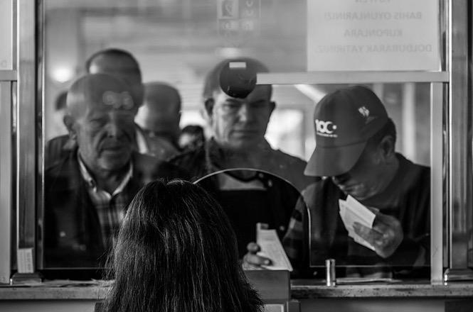Foto horizontal: pessoas apresentam documentos em guichê de atendimento, em imagem em preto e branco.