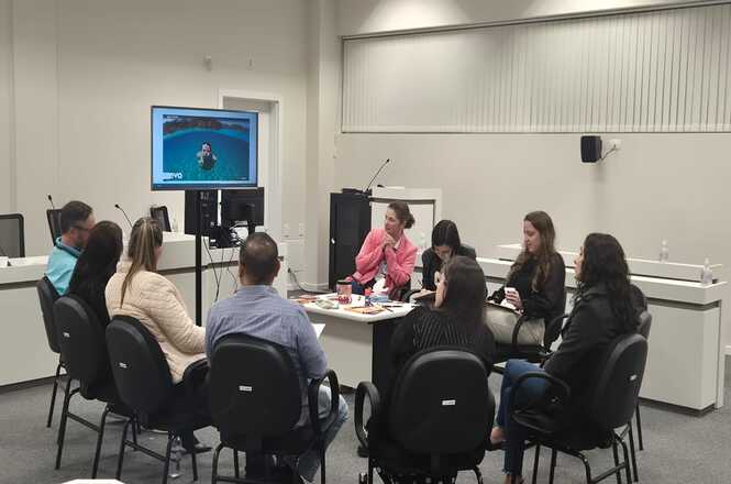 Foto horizontal: participantes realizam atividade em grupo ao redor de mesa com materiais educativos, enquanto acompanham conteúdo exibido em tela.