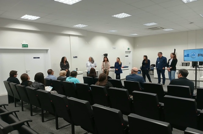 Foto horizontal: equipe apresenta atividade diante de participantes reunidos em auditório, com tela de projeção ao lado.