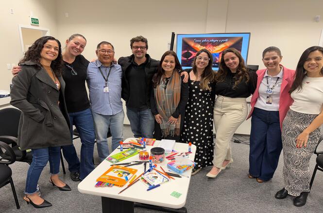 Foto horizontal: grupo de participantes posa para foto após atividade, diante de mesa com materiais educativos, livros, lápis e objetos coloridos.