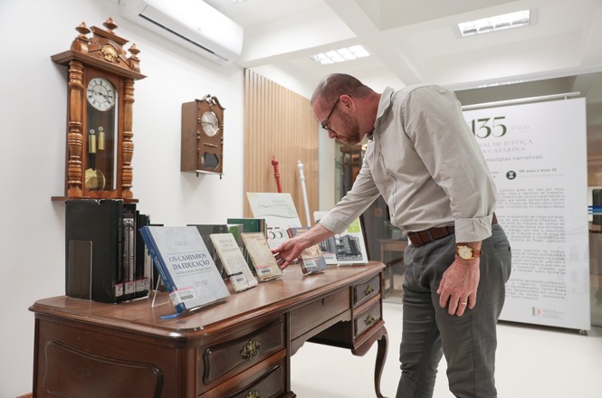 Foto horizontal: homem em sala branca observa livros expostos sobre mesa de madeira antiga em mostra.