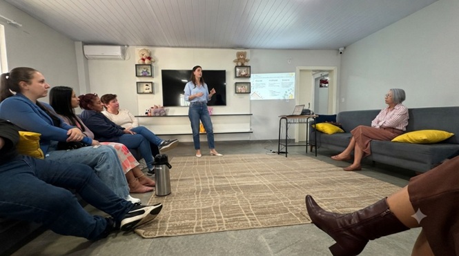 Foto horizontal: mulher conduz apresentação para participantes sentados em sala de atendimento.