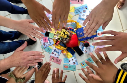 Várias mãos estendidas em círculo ao redor de um conjunto de objetos e cartões coloridos dispostos no chão, indicando uma dinâmica em grupo.