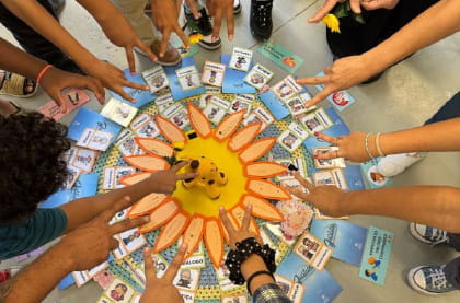 Várias mãos de pessoas apontando para um jogo circular colorido, com um sol no centro e cartas com imagens ao redor.