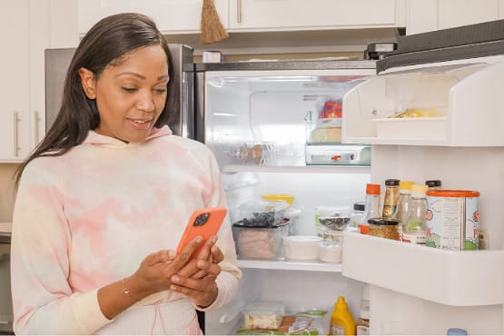 mulher olhando para o celular na frente de geladeira aberta