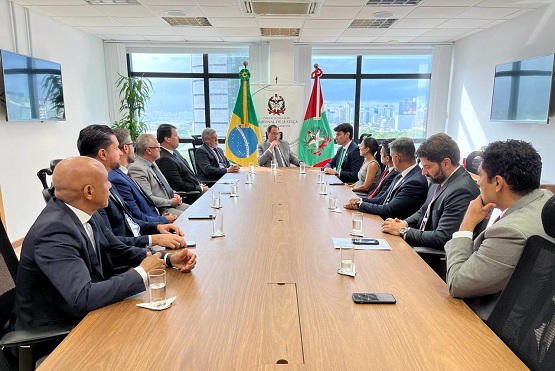 Foto horizontal: entre as bandeiras do Brasil e de SC, Francisco Oliveira Neto conduz reunião com autoridades, tons claros.