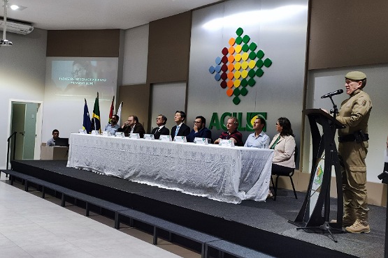 A foto parece ser de um evento ou conferência, onde várias pessoas estão sentadas em uma mesa longa coberta com uma toalha branca. No púlpito, também no palco, um homem com farda policial está falando. Há um painel de fundo com um logotipo colorido composto por várias esferas dispostas em um padrão triangular.