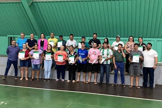Uma foto de um grupo de pessoas posando para a câmera, todas estão lado a lado segurando certificados. Eles estão em um ambiente fechado, provavelmente um ginásio, o teto é verde e o piso tem linhas.