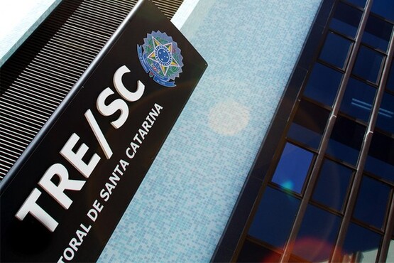 A imagem mostra uma construção, que parece ser alta, com muitos vidros azuis na fachada. Bem na frente da foto há uma placa na vertical escrito "TRE-SC".
