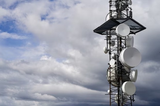 A imagem mostra o topo de uma torre de telecomunicações no fundo o céu está azul com várias nuvens.