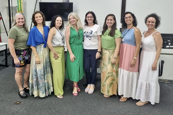 A imagem mostra um grupo de oito pessoas, todas são mulheres posando para uma foto. Elas estão em pé, lado a lado, em um ambiente interno. Estão vestidas com roupas coloridas e variadas, incluindo vestidos longos e saias. Todas estão sorrindo.