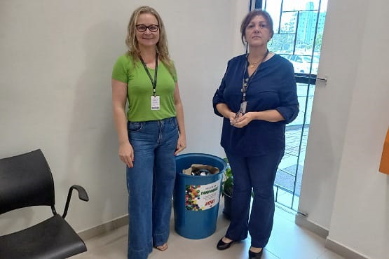 A imagem mostra duas mulheres em pé, uma ao lado da outra, em um ambiente interno. A mulher à esquerda está vestindo uma camiseta verde e calça jeans azul, e usa um crachá pendurado no pescoço. A mulher à direita está vestindo um uniforme azul escuro. Entre elas, há um recipiente azul com uma abertura na parte superior, que parece ser um coletor de tampinhas plásticas. Ao fundo, há uma cadeira preta e uma parede clara.