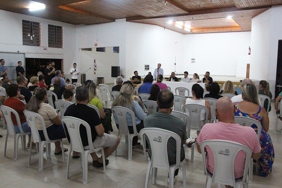 A imagem mostra uma reunião ou evento em um salão fechado. As pessoas estão sentadas em cadeiras de plástico branco, voltadas para a frente, onde parece haver uma apresentação ou discurso acontecendo. A maioria das pessoas está vestida de forma casual. No fundo, há uma pessoa em pé, possivelmente falando ao público.