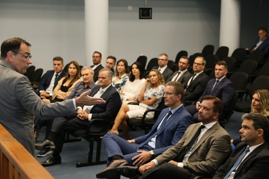 A imagem mostra uma reunião ou conferência com várias pessoas sentadas em cadeiras, prestando atenção em um homem que está de pé, falando e gesticulando com as mãos. As pessoas na audiência estão vestidas formalmente, sugerindo um ambiente profissional.