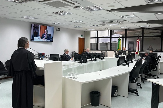 A imagem mostra uma sala de reuniões ou tribunal. Há várias pessoas sentadas em torno de uma mesa grande e branca, usando ternos. Na parede ao fundo, há uma televisão que exibe a imagem de uma pessoa falando. Também é possível ver bandeiras do Brasil e de outro estado ou município ao fundo, perto de uma porta.