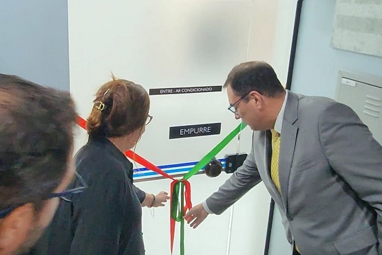 A foto que parece ser de um evento de inauguração ou corte de fita. Na foto, há duas pessoas: uma mulher de costas e um homem de perfil, ambos segurando fitas coloridas (vermelha, verde e azul). O homem está vestindo um terno cinza e uma gravata amarela.