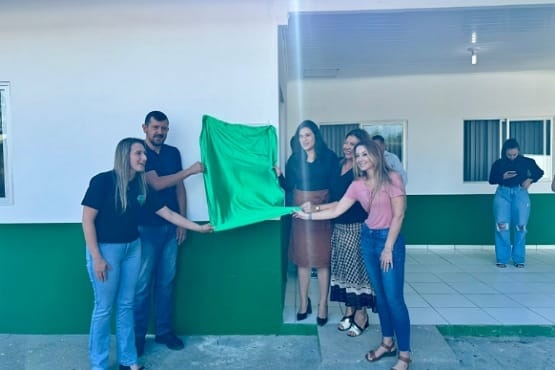 Na foto, há um grupo de pessoas posando juntas, são 4 mulheres e 1 homem. Todos estão segurando um pano verde que está próximo a uma parede, em um ambiente fechado.  No fundo há janelas e mais algumas pessoas.