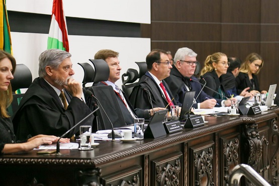 A imagem mostra um grupo de pessoas sentadas em uma mesa, aparentemente em uma reunião formal ou sessão de tribunal. As pessoas estão vestidas de forma formal, com ternos e togas. Há microfones na mesa à frente deles. Ao fundo, é possível ver bandeiras, incluindo a bandeira do Brasil. A imagem está inclinada para a direita.