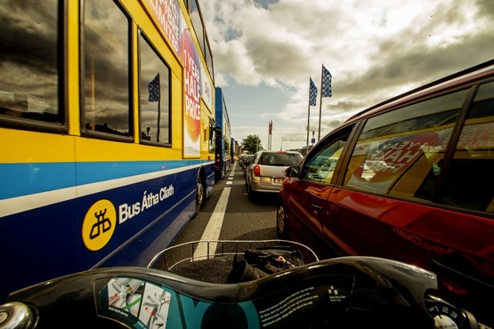 A imagem mostra a visão de quem está dirigindo uma moto. O motorista está ultrapassando o trânsito entre ônibus e carro. Na frente, ao longo da estrada há outros carros e placas de trânsito. O sol está entre as nuvens, mas mesmo assim ilumina.