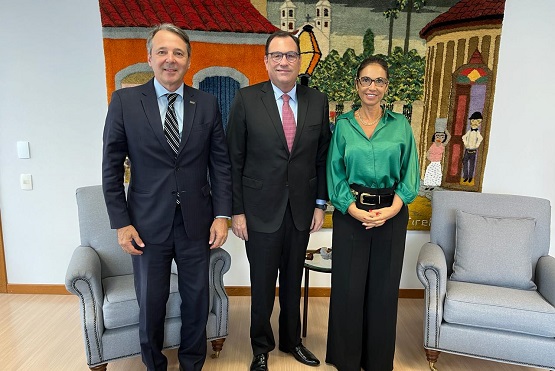 Três pessoas estão em pé, lado a lado,  posando para a foto, são dois homens e uma mulher. Eles estão sorrindo. Atrás deles há um painel colorido com a imagem de uma casa e algumas pessoas, parece ser feito com fio. De cada lado da foto há uma poltrona cinza, que parece confortável.