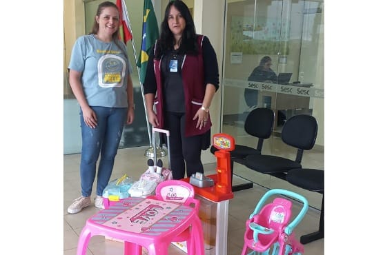 A imagem mostra duas mulheres em pé em um ambiente interno. A mulher à esquerda está vestindo uma camiseta cinza e calça jeans, enquanto a mulher à direita está usando um colete bordô sobre uma blusa preta e calça preta. Atrás delas, há uma bandeira do Brasil e uma bandeira do estado de Santa Catarina. No chão, à frente das mulheres, há uma mesa infantil rosa com cadeiras combinando, um carrinho de bebê de brinquedo e uma caixa registradora de brinquedo.