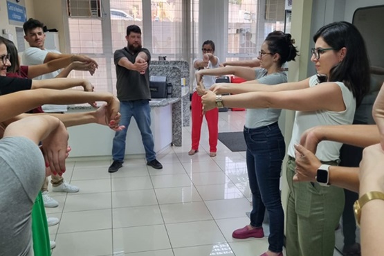 A imagem mostra um grupo de pessoas em um ambiente interno realizando exercícios de alongamento. Elas estão em pé, formando um círculo, com os braços estendidos à frente e as mãos dobradas para baixo, segurando os dedos com a outra mão. Ao fundo, há uma bancada com alguns objetos e janelas que permitem a entrada de luz natural. As pessoas parecem estar concentradas na atividade.
