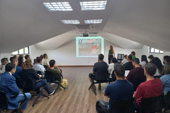A imagem mostra um grupo de pessoas sentadas assistindo uma mulher ruiva falar na frente de todos. Um telão exibe uma projeção de uma imagem colorida. O ambiente é fechado, o teto é branco e faz formato de triângulo, o chão é de madeira.