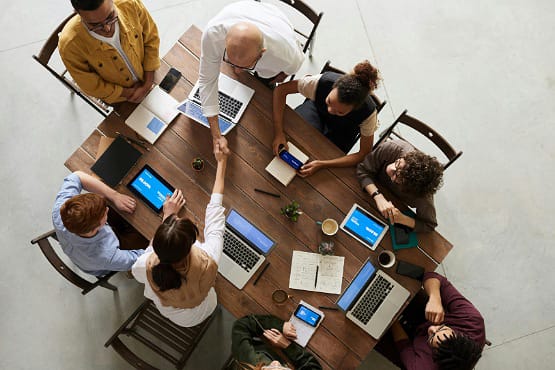 A foto foi tirada de cima para baixo, aparecendo o tampo de uma mesa de madeira com seis pessoas sentadas em sua volta. Elas estão com notebooks, papéis e canetas. Aparentemente estão fazendo um trabalho em grupo.
