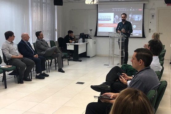 A imagem mostra alguns homens e mulheres sentados em cadeiras verdes formando um corredor, dentro de uma sala fechada Um homem mais a frente está em um púlpito discursando com um telão nas suas costas com imagens coloridas.