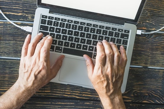 As mãos de uma pessoa estão sobre um teclado de notebook, que está em cima de uma mesa de madeira. Alguns cabos como carregador e fone estão conectados no notebook.
