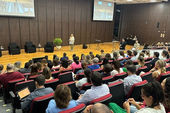 A imagem mostra um auditório cheio de pessoas assistindo a uma apresentação. A palestrante está em pé no palco, atrás de um púlpito, falando para a audiência. O auditório tem cadeiras vermelhas e pretas, e as pessoas estão sentadas, prestando atenção. No palco, há três cadeiras pretas e uma mesa pequena com um vaso de planta. Duas telas grandes estão penduradas na parede de madeira escura atrás da palestrante, exibindo a mesma imagem. Há também alguns equipamentos de gravação e pessoas trabalhando ao fundo, possivelmente registrando o evento.