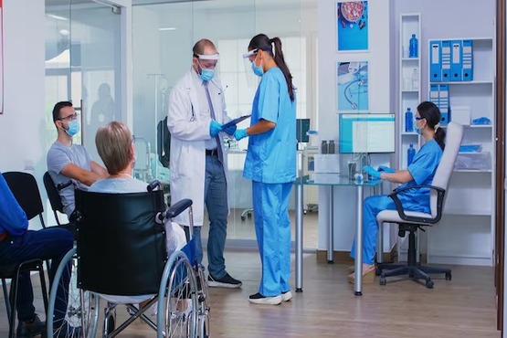 Uma sala de hospital em tons azuis e brancos. Dois pacientes estão esperando, um usa cadeira de rodas e o outro está de máscara. Dois médicos estão de pé no meio da imagem, um homem e uma mulher, ele está segurando uma prancheta e ela está prestando atenção. Mais no canto da imagem uma terceira médica está sentada em uma mesa de vidro com um computador em sua frente
