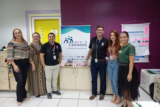 Seis pessoas estão posando para uma foto, sendo 2 homens e 4 mulheres. Todos estão sorrindo, três estão lado a lado na direita da imagem e os outros três lado a lado na esquerda, um balcão pequeno de duas portas está dividindo o grupo. Eles estão em uma sala fechada e no fundo há uma parede bordo com dois banners pendurados.