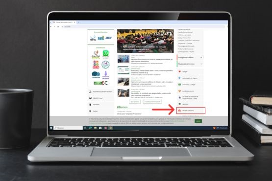 Um computador está em cima de uma mesa, do lado há uma caneca e alguns livros. A tela do computador está aberta do site do TJSC e uma flecha vermelha está mostrando onde fica o Plantão Jurídico no site.