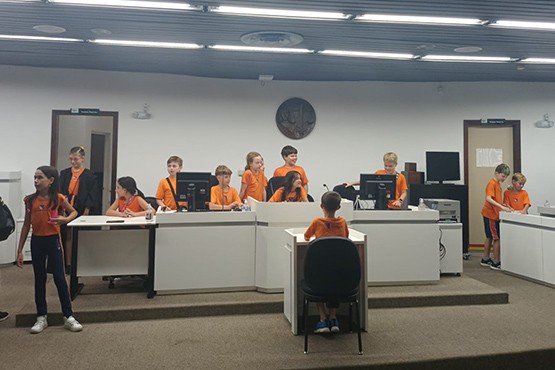 A imagem mostra várias crianças com uniformes laranjas e azuis em uma sala fechada. Há uma mesa branca comprida onde eles estão brincando, mais atrás tem duas portas que estão abertas. O ambiente parece ser uma sala de audiência.