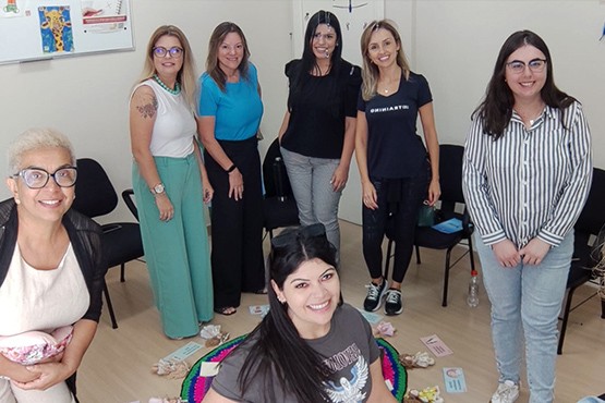 A imagem mostra 7 mulheres posando para a foto, todas elas estão sorrindo. Elas estão em uma sala fechada e ao fundo estão posicionadas algumas cadeiras pretas.